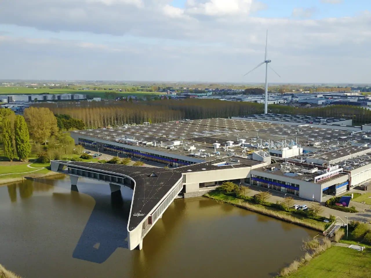 A large industrial building extends over a waterway, featuring a unique overhanging structure. Surrounding areas include green fields and a wind turbine, with industrial facilities in the distance.