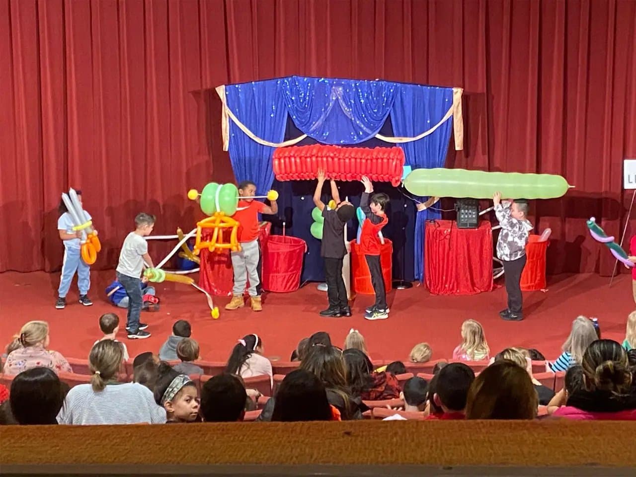 Children on stage manipulate large balloon structures in a theater setting, with an audience of seated spectators. A blue and red backdrop, with red curtains, decorates the stage.