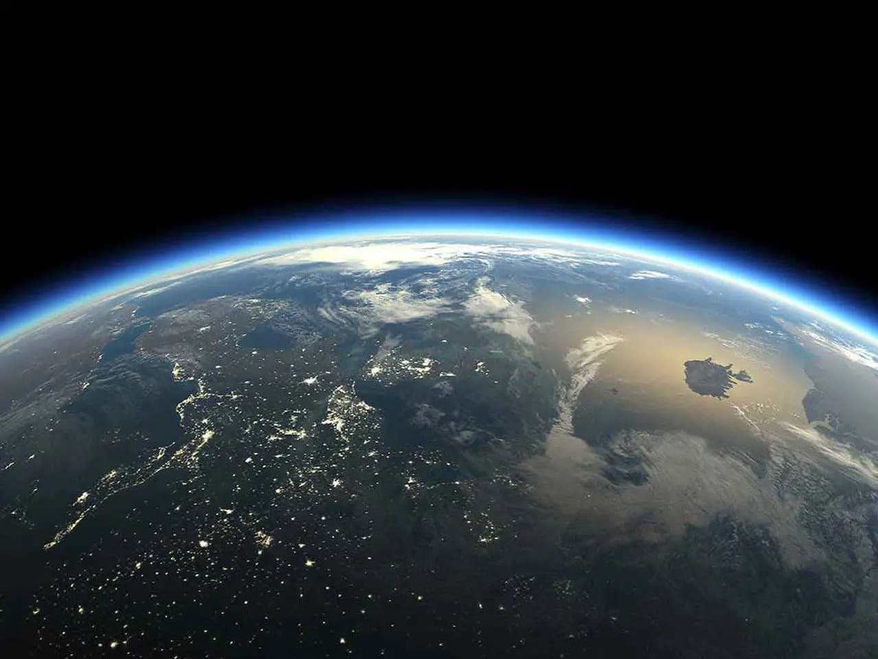 Earth viewed from space, partly illuminated with visible city lights. Wispy clouds float over landmasses, and a blue atmosphere contrasts with the dark expanse of space above.