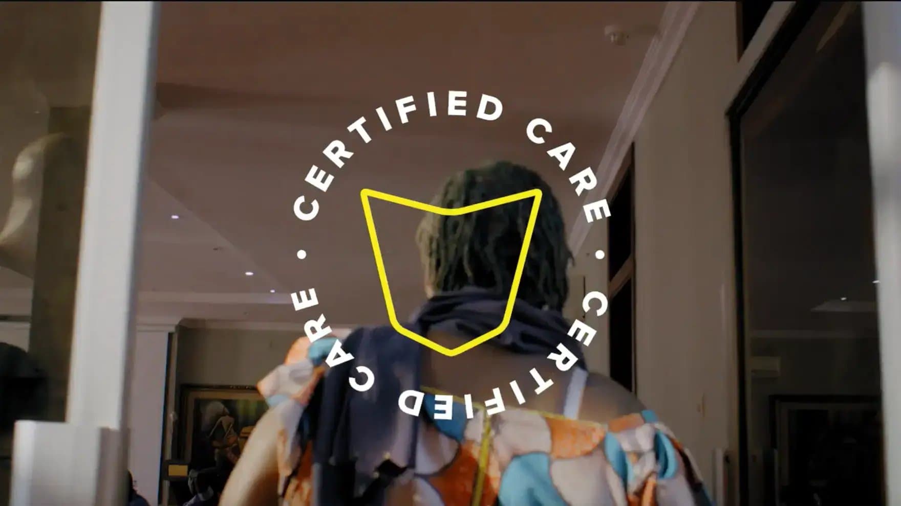 A person wearing colorful clothing enters a room, with "CERTIFIED CARE" text encircling a yellow outline overlaid on the image. The setting is indoors, with modern decor.