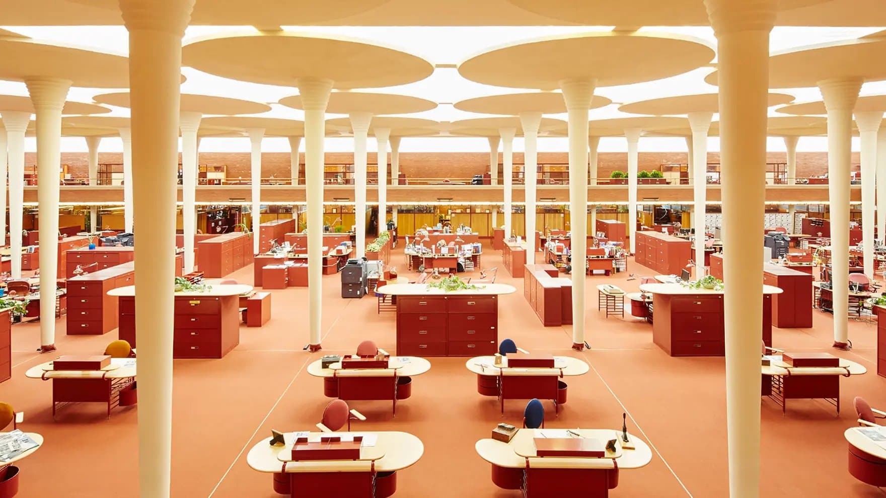 Desks are arranged in a spacious open-plan office, surrounded by red cabinets and supported by white columns beneath a uniquely patterned ceiling. The environment is neat and organized.