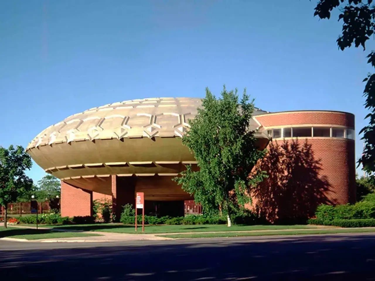A large, dome-shaped building with a unique, geometric roof design sits among trees and grass. The structure is made of red brick, and its modern style stands out in a sunny setting.