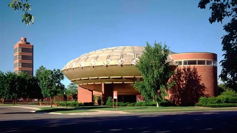 Dome-shaped building with a waffle-like structure sits elevated on brick supports, surrounded by trees and greenery. A cylindrical tower is visible in the background, under a clear blue sky.
