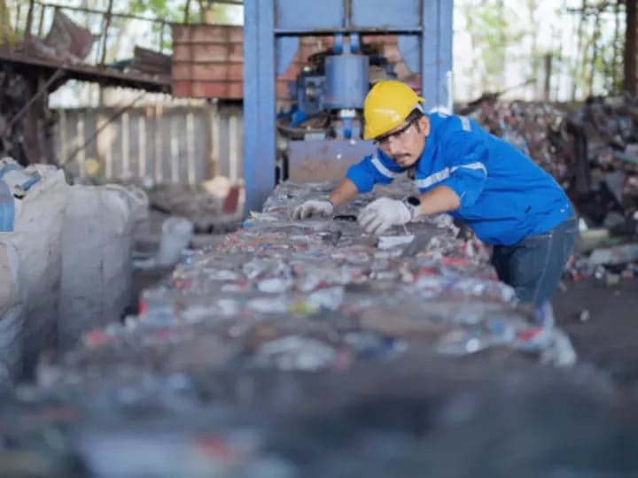 Worker wearing a yellow helmet and blue uniform inspects materials on a conveyor belt in an industrial recycling facility, surrounded by bags and machinery.