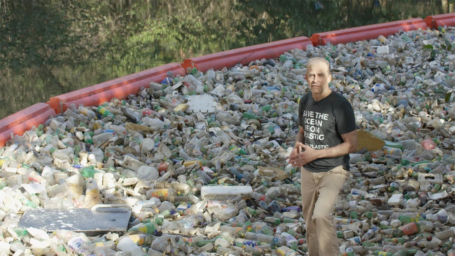 A person stands amid a dense collection of plastic waste enclosed by orange barriers. Their shirt reads "SAVE THE OCEAN FROM PLASTIC #NOMOREPLASTIC," highlighting a polluted environmental issue near a water body.