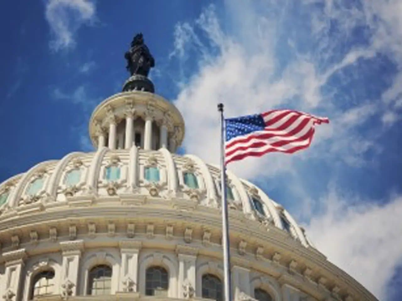 A U.S. flag waves above the dome of a classical capitol building. The scene is set against a clear, blue sky with scattered clouds.