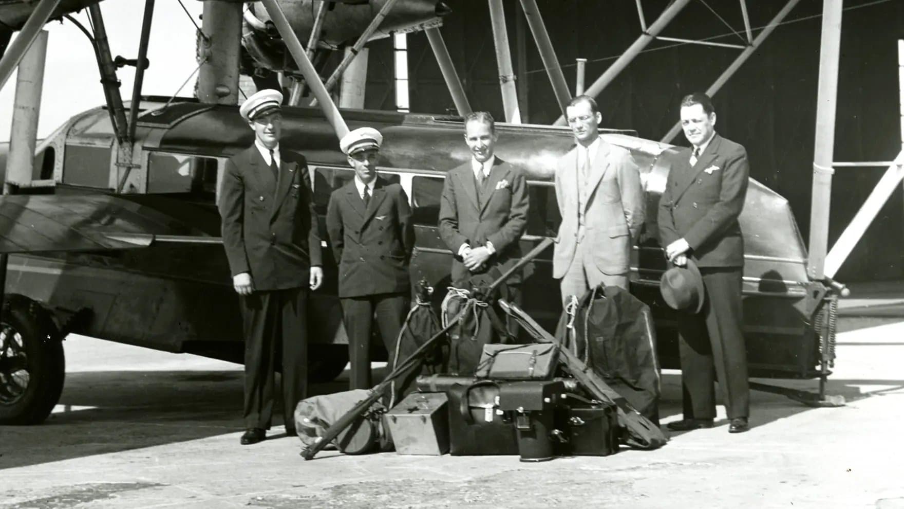 Five men stand in front of a vintage airplane, dressed in suits and uniforms, with luggage and equipment placed on the ground. The setting is an airport hangar.