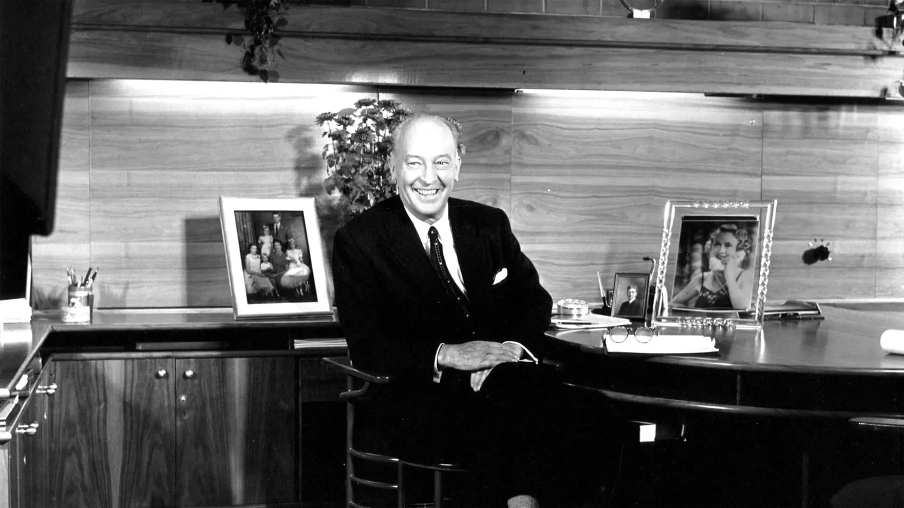 A man sits smiling at a desk with framed family photos, glasses, and a notepad, in an office featuring wood paneling and a potted plant.
