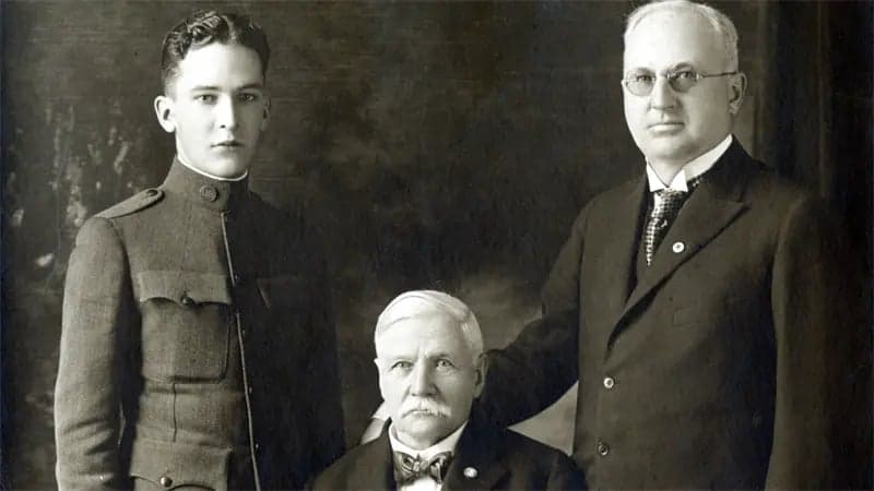 Three men pose formally; one in military uniform, another seated with a serious expression, and the third wearing glasses and a suit. They are in a neutral, indoor setting.