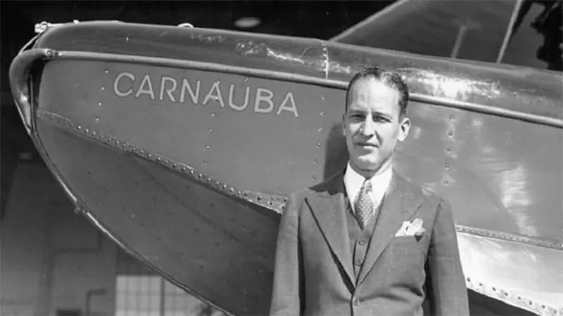 A man in a suit stands in front of an airplane with the name "CARNAÚBA" visible on its fuselage. The scene is outdoors with an industrial background.