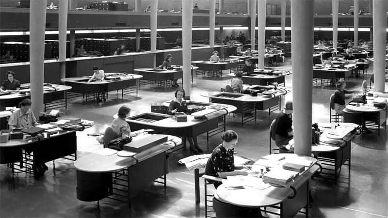 Workers are seated at large wooden desks, typing and organizing papers in a spacious, open-plan office with high ceilings and numerous vertical columns, suggesting a mid-20th-century business setting.