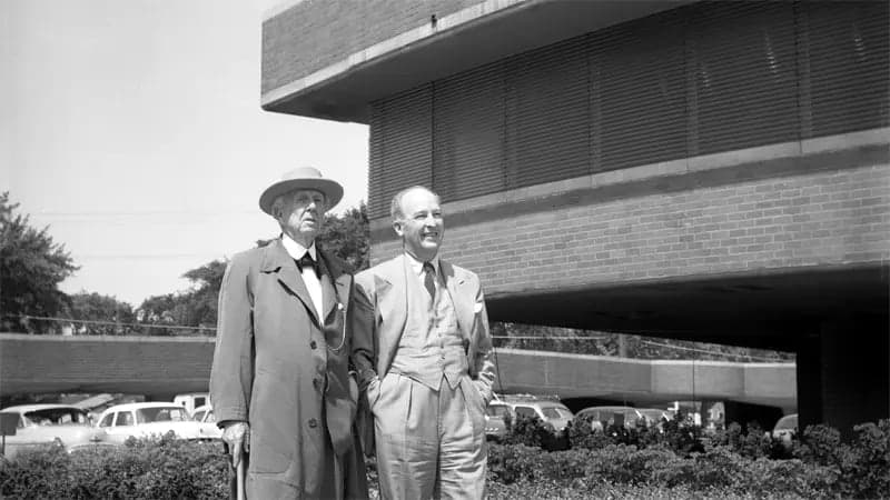 Two men in suits stand side by side outside a modern brick building. They appear to be observing something. Cars and trees are visible in the background, creating an urban setting.