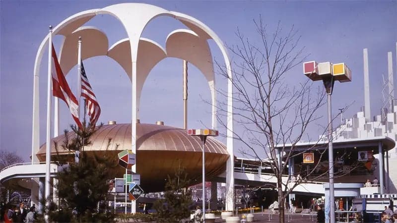 A futuristic pavilion with a domed structure is encircled by tall, arching white beams. Flags flutter nearby. Sparse trees and a partly cloudy sky complete this mid-20th-century exhibition setting.