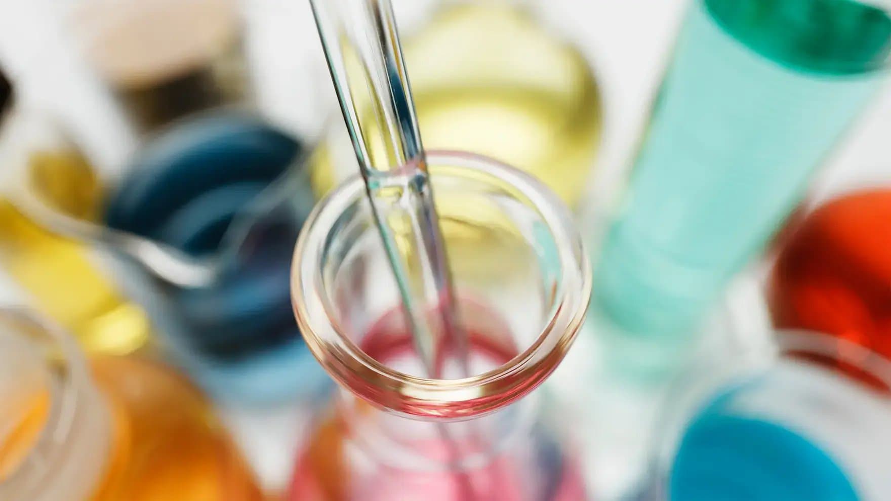 A glass pipette dips into a clear flask containing pink liquid, surrounded by various colored liquids in blurred laboratory beakers.