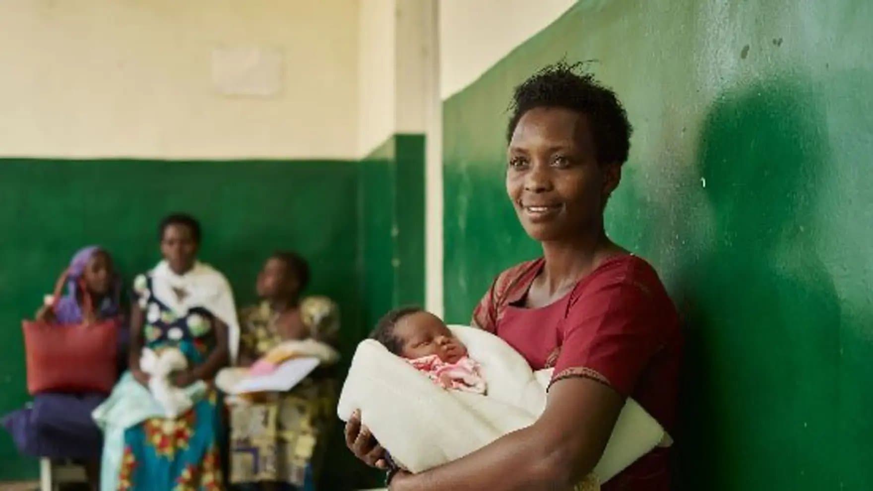 A woman in a maroon dress holds a sleeping baby wrapped in a white blanket, standing against a green wall. In the background, several women are seated.
