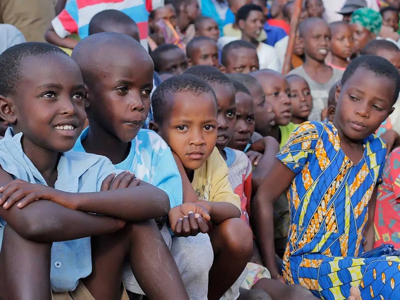 Children are sitting closely together outdoors, appearing attentive and engaged. They are surrounded by a larger group of people, suggesting a communal gathering or event.