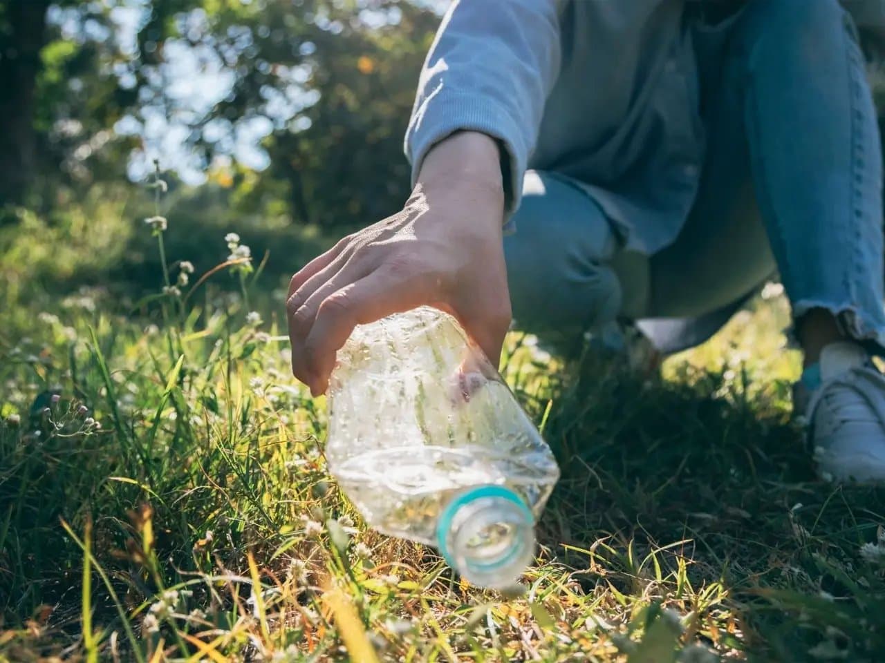 A hand picks up a plastic bottle from grass, set in a sunny outdoor environment with greenery and soft daylight filtering through trees.