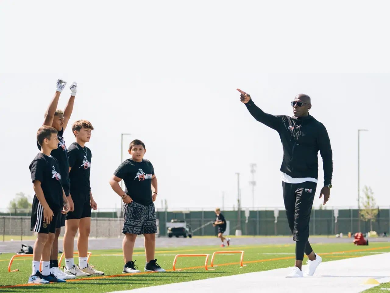 A coach instructs four children on a sports field, pointing in guidance. The kids, in athletic gear, appear attentive. Orange hurdles are arranged on the green turf.