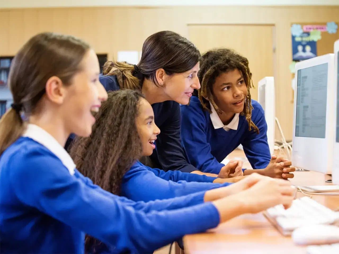 Students wearing blue uniforms use computers enthusiastically in a classroom setting, accompanied by a teacher who engages with them, smiling, against a backdrop of wooden walls and bulletin boards.
