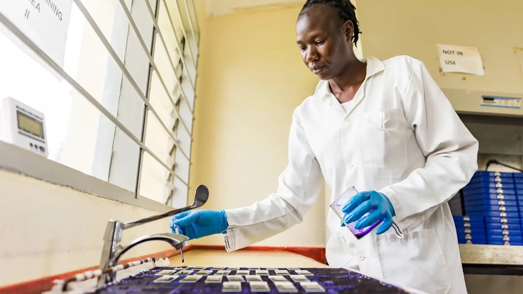A person in a lab coat and gloves uses a dropper and holds a beaker over a table with glass slides. Sign reads "NOT IN USE," with a window nearby.