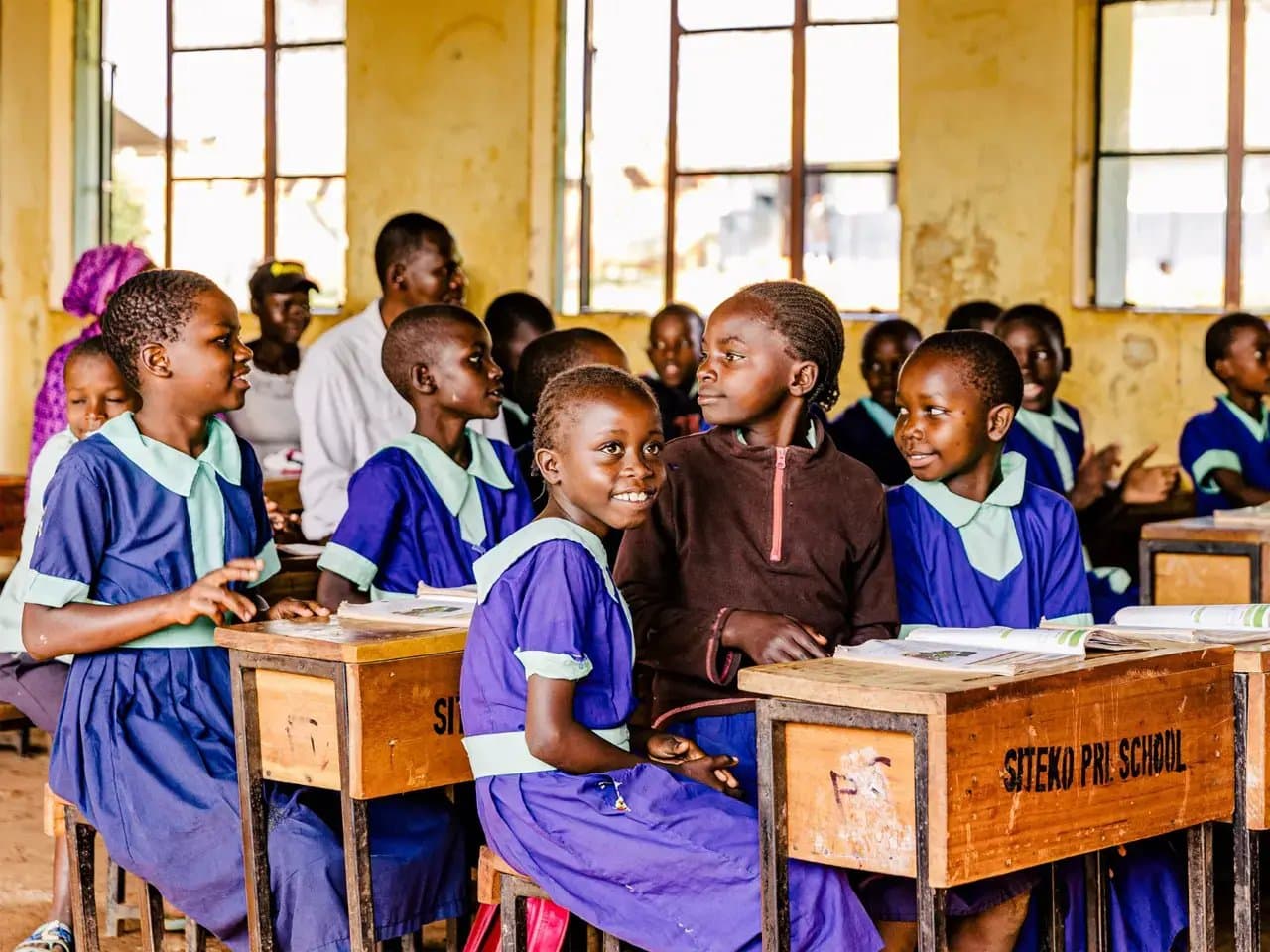 Children in blue uniforms sit and interact at wooden desks labeled "Sitekø Pri. School" in a classroom with large windows, surrounded by more students and a few adults.
