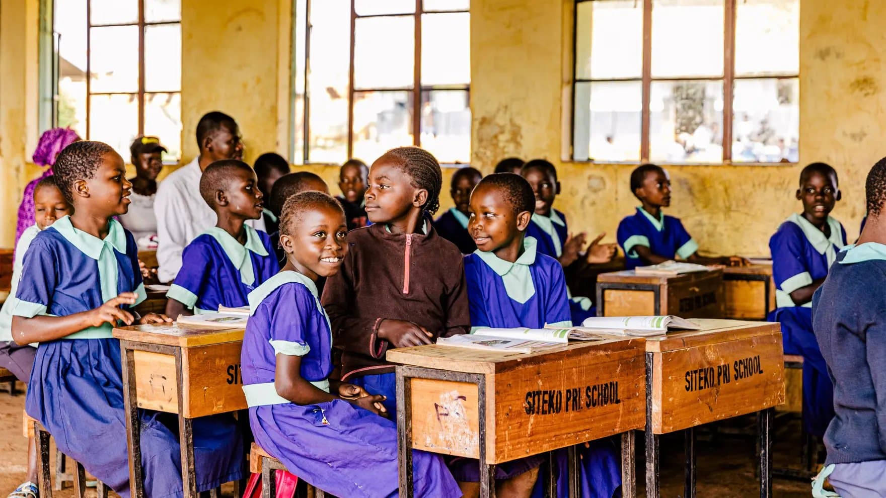 Children in blue uniforms sit at wooden desks, some smiling, in a classroom with yellow walls. Two desks feature the text "SITEKO PRI SCHOOL." Sunlit windows line the wall.