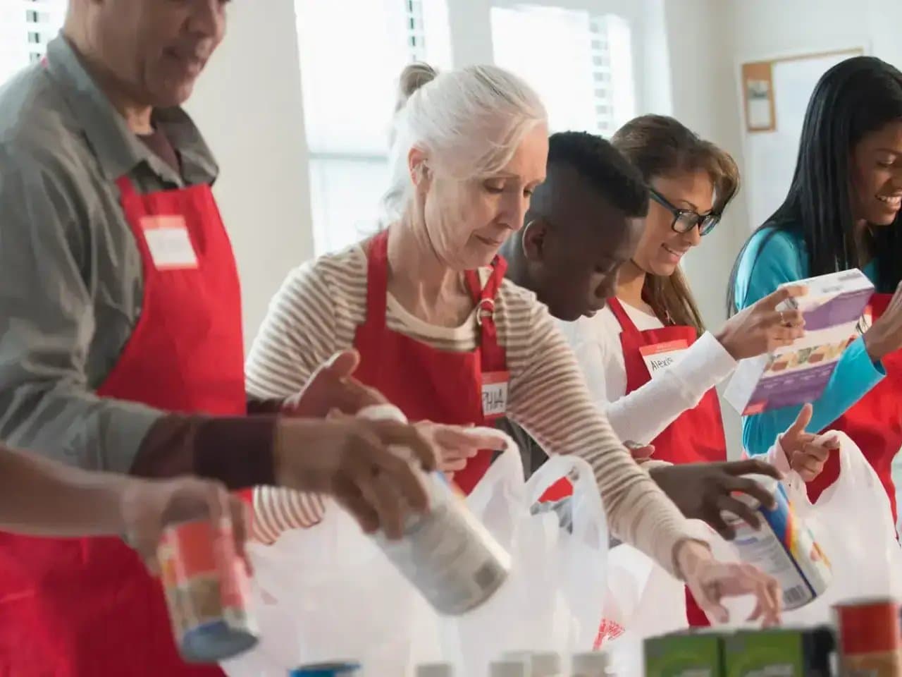People wearing red aprons pack food into bags, working together in a bright room with windows. Name tags are visible, including "Alexis." They focus intently on organizing the items.