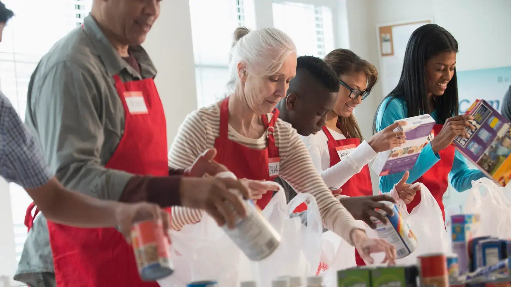 Volunteers wearing red aprons organize canned goods and food items into plastic bags. They work together in a well-lit room with large windows, focusing on community service tasks.