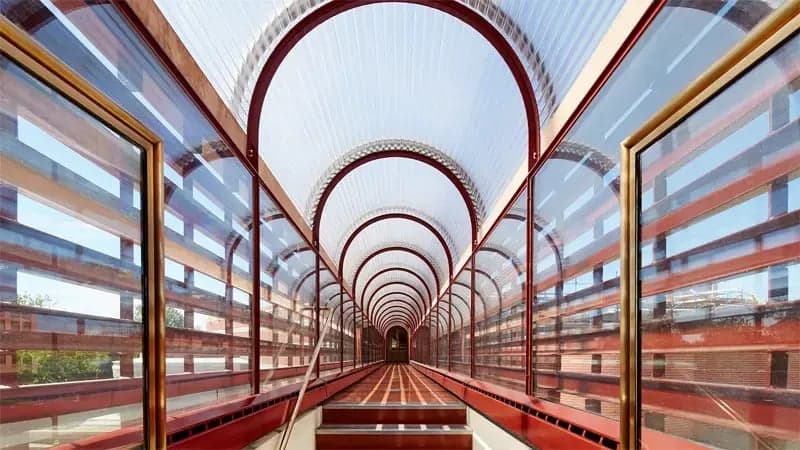 A long, enclosed walkway arches overhead with transparent, curved roof panels, bordered by red frames. The pathway extends straight, surrounded by clear side panels offering an urban view.