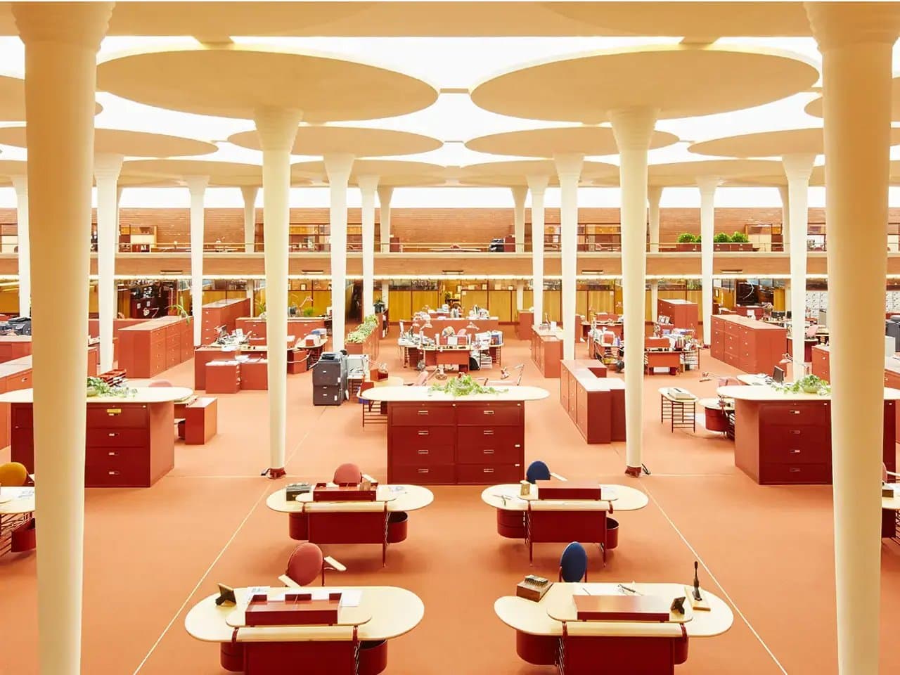 Desks with red cabinets are arranged in an expansive, open-plan office. Circular columns and plant-topped dividers create a structured but airy environment. The space is brightly lit.