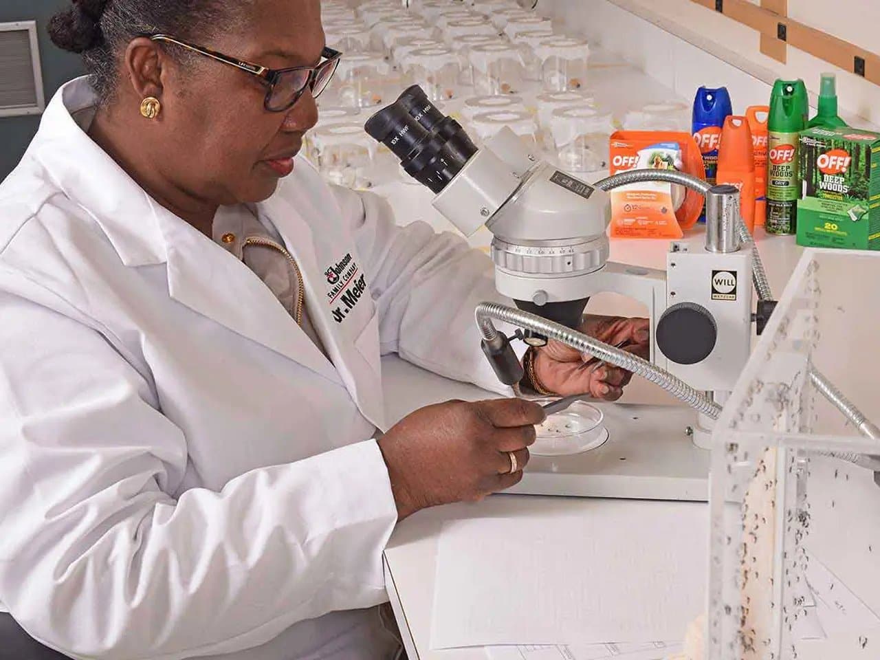 A scientist examines a specimen under a microscope in a lab. The table holds various insect repellent products. She wears a lab coat labeled "Dr. Melear."