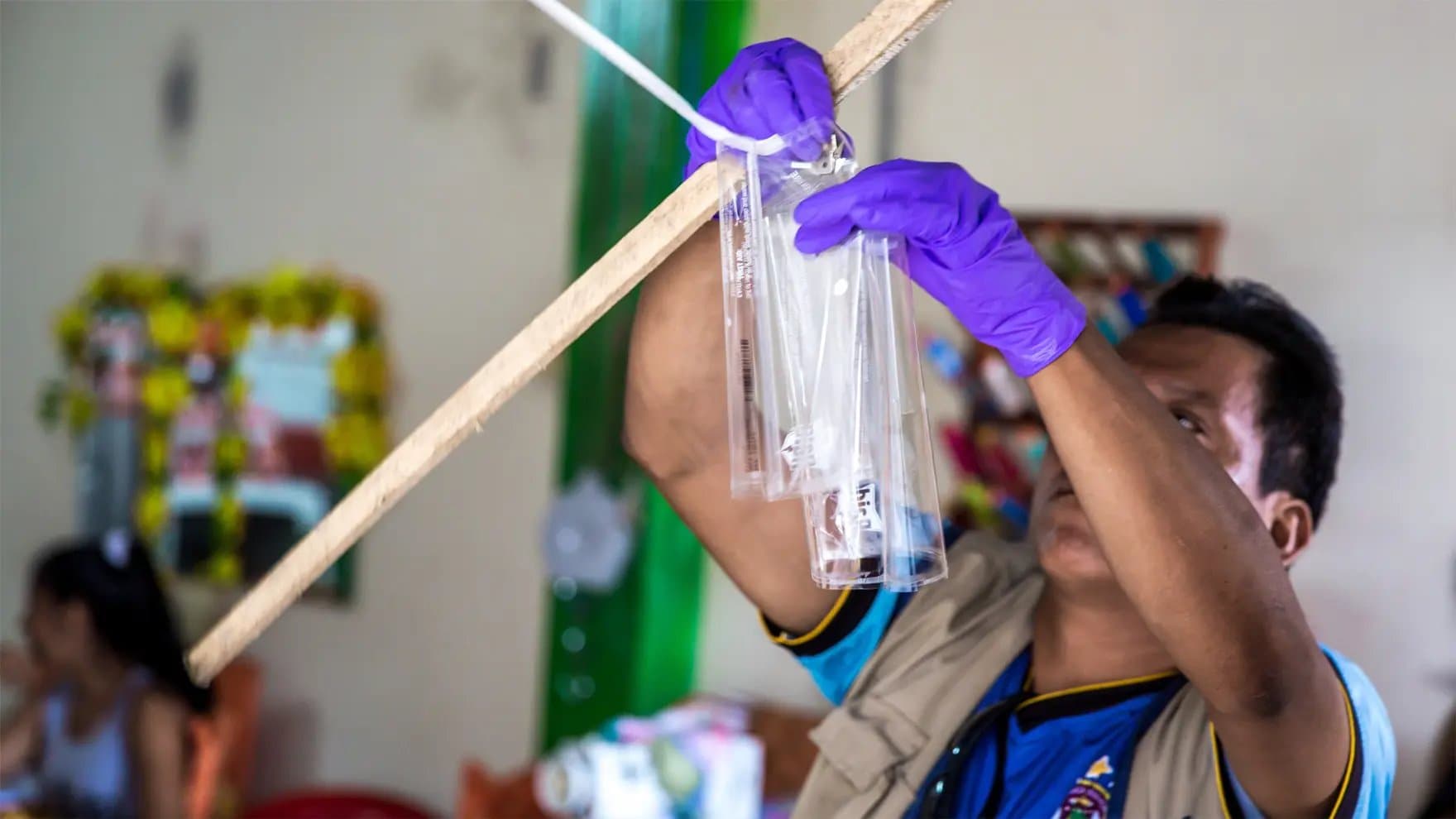 A person in purple gloves secures transparent bags to a wooden stick, possibly preparing a setup, in an indoor room with blurred decorations and seated people in the background.