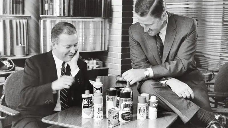 Two men in suits are sitting at a table, examining various household products, including "Klear," "Raid," "Glade," "Pledge," and "Oust," in an office filled with bookshelves.