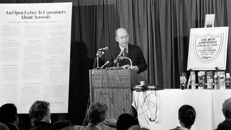 A man stands at a podium speaking into multiple microphones during a press conference. Behind him, a large poster reads "An Open Letter to Consumers About Aerosols," and nearby, another sign displays "USE WITH CONFIDENCE: CONTAINS NO FREON OR OTHER FLUOROCARBONS KNOWN TO HARM THE ENVIRONMENT." A row of aerosol cans is visible on a table. An audience listens in the foreground.
