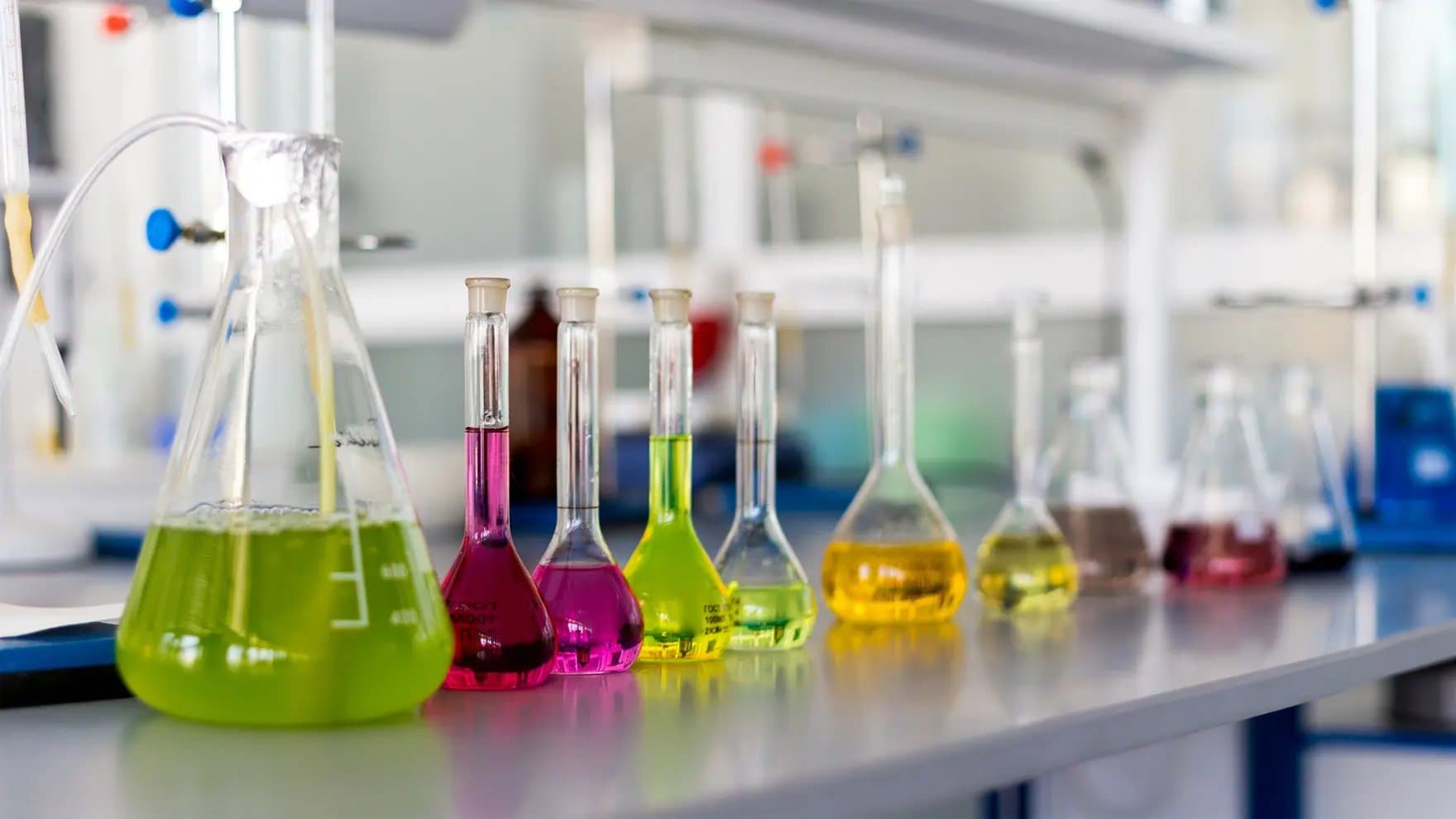 Flasks contain colorful liquids arranged in a line on a laboratory bench. Surrounding scientific equipment suggests an active research environment.