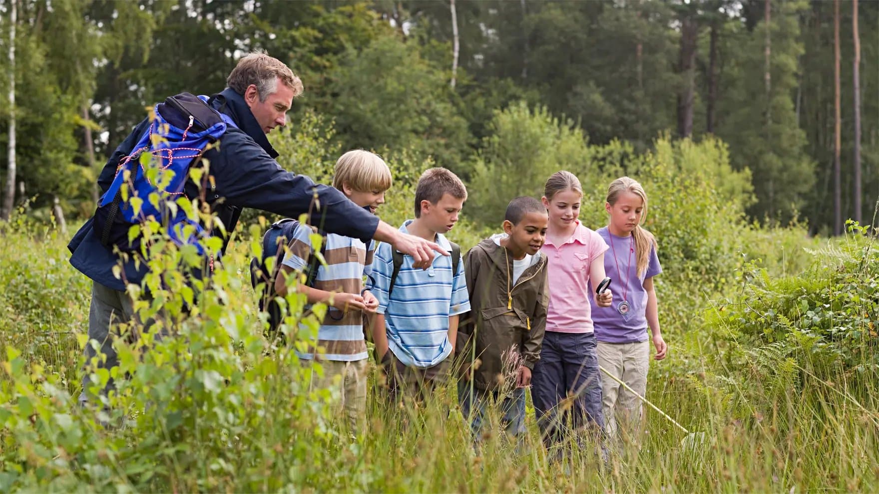 A group of children and a teacher are observing nature in a lush forest, surrounded by greenery, as the teacher points at something interesting in the grass.