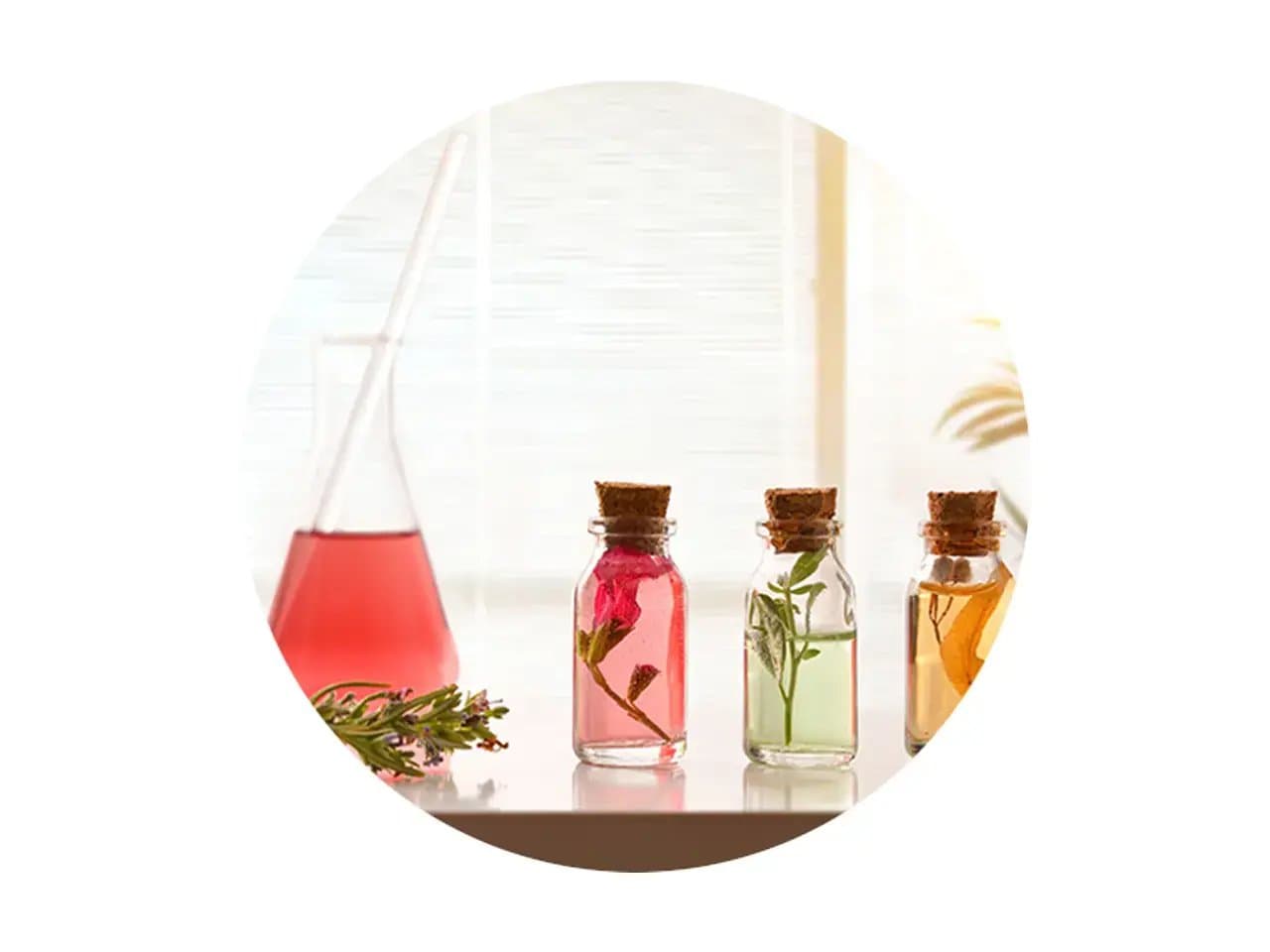 Glass bottles with cork stoppers contain colorful liquids and herbs, sitting on a white table. In the background, a flask with a pink liquid and sprigs of greenery are visible.