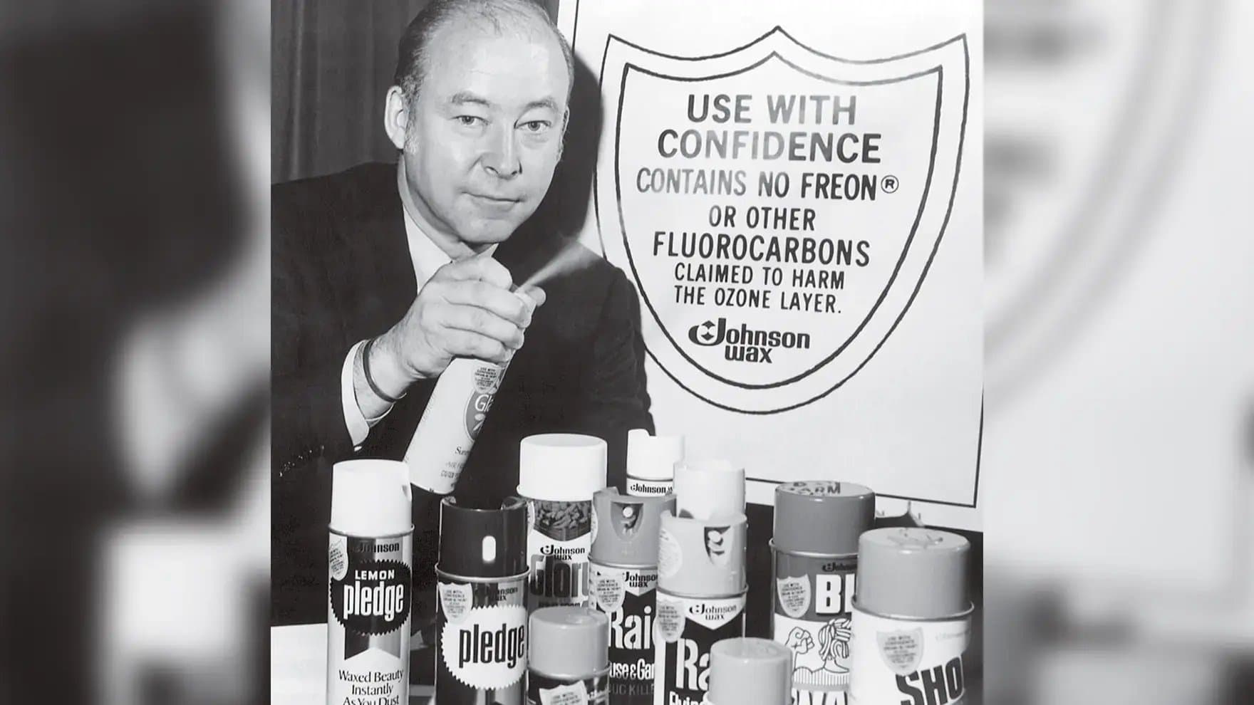 A person sprays an aerosol can, surrounded by various cleaning products. Behind him, a sign reads: "USE WITH CONFIDENCE CONTAINS NO FREON OR OTHER FLUOROCARBONS CLAIMED TO HARM THE OZONE LAYER. Johnson Wax."