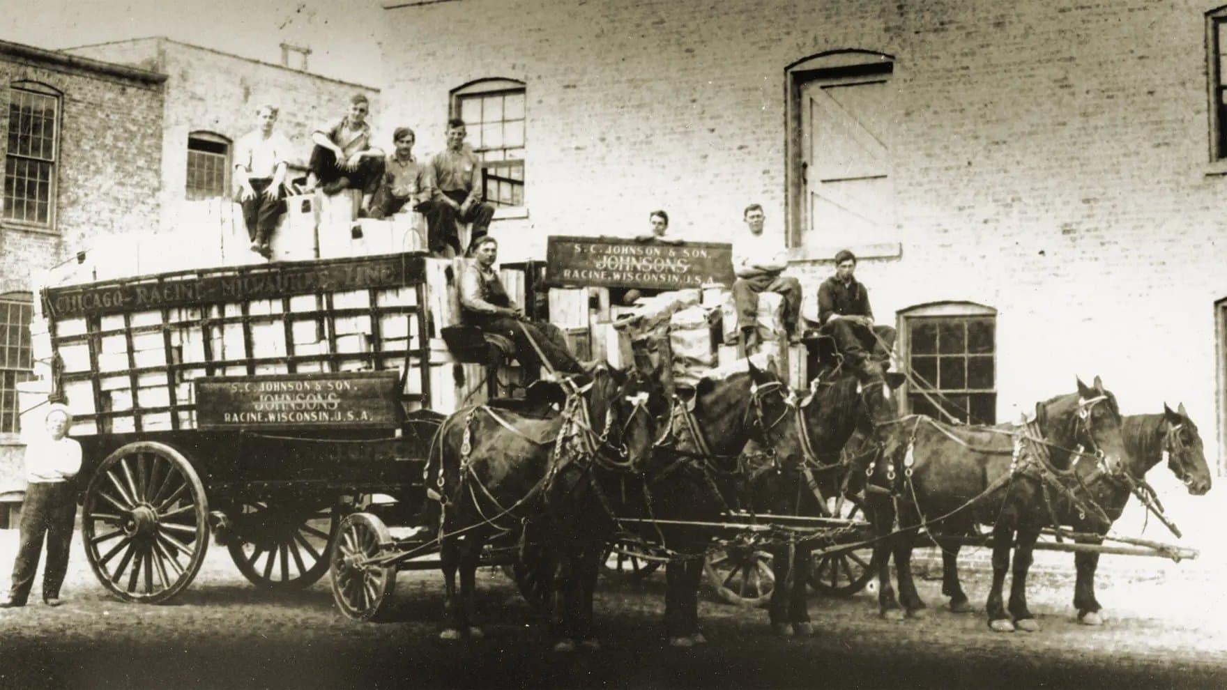 A large horse-drawn wagon, loaded with boxes and several men seated on top, stands in front of an industrial building. The wagon displays the text: "S.C. JOHNSON & SON, JOHNSON’S, RACINE, WISCONSIN, U.S.A."