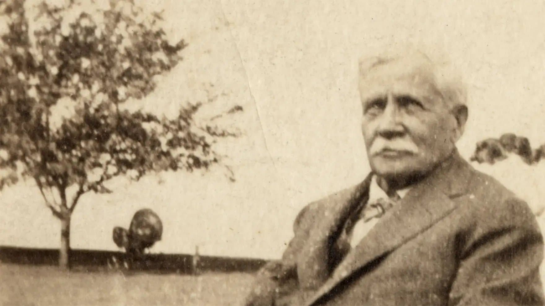 A man with a mustache sits in a suit outdoors, beside a blurred tree and distant trees in the background.