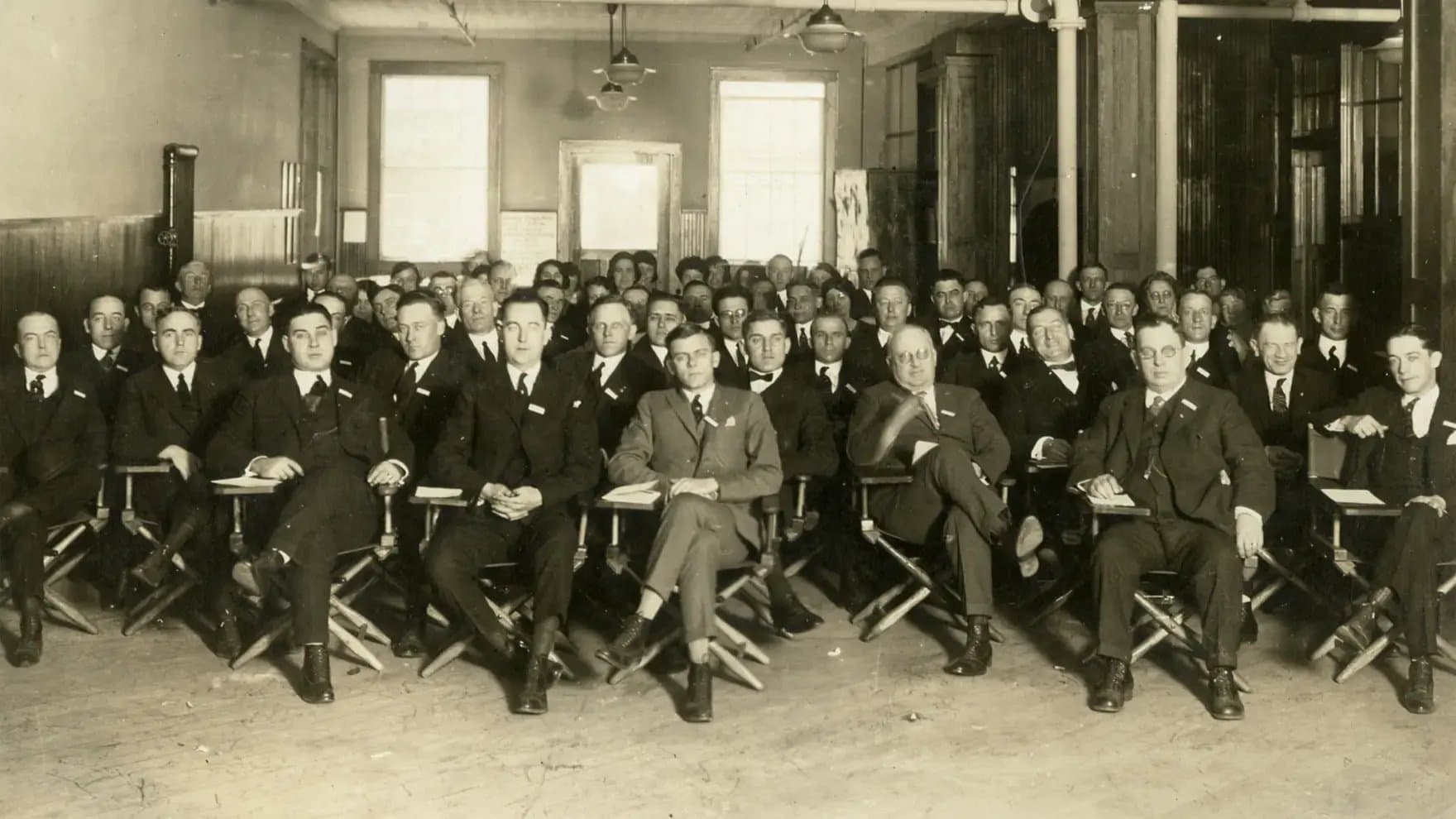 A large group of men in suits sit on folding chairs, attentively facing forward. The setting is a vintage indoor room with wooden floors and visible windows.