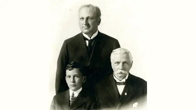 Three men in formal attire pose together; a young boy sits between two older men in suits, set against a plain background. The image is black and white.
