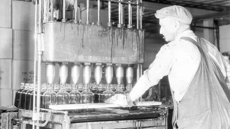 Worker operates a bottle-filling machine in an industrial setting. Glass bottles align beneath the dispenser, while cardboard boxes stack in the background. The worker wears overalls and a cap.