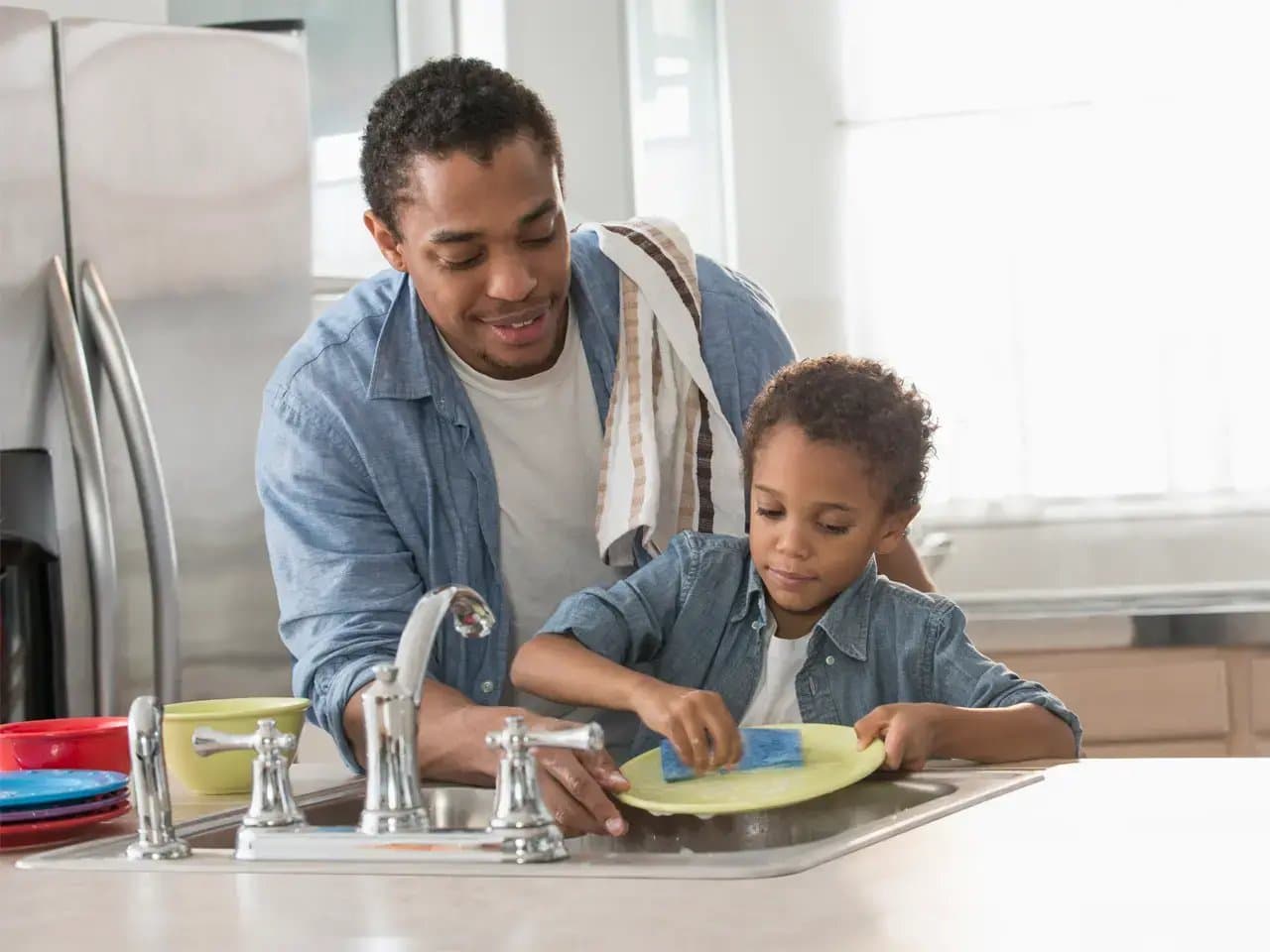 A child cleans a plate with a sponge at the kitchen sink while an adult, draped with a towel, supervises. Nearby, colorful bowls create a lively atmosphere.