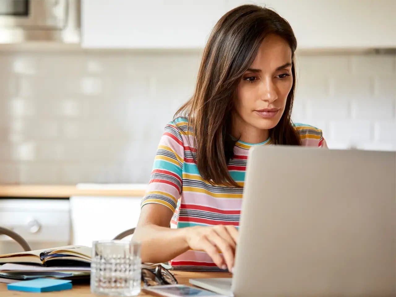 A person is focused on using a laptop in a kitchen setting. Nearby are an open notebook, glass of water, and phone on the table.