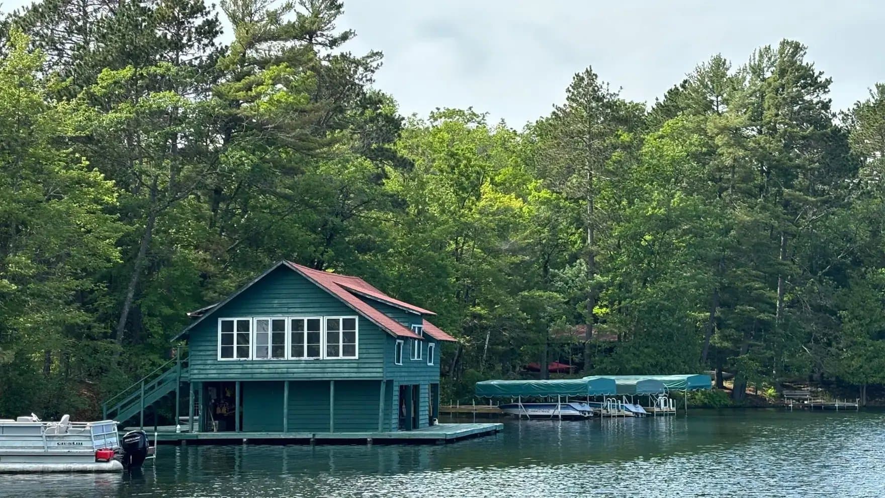 A green boathouse with windows stands on a lake, surrounded by lush trees. Nearby, boats are docked under a covered area, enhancing the tranquil setting.