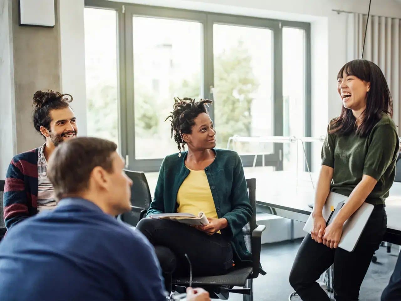 A group of four people are sitting together, engaged in conversation, with one holding a laptop. They are in a bright room with large windows and chairs.