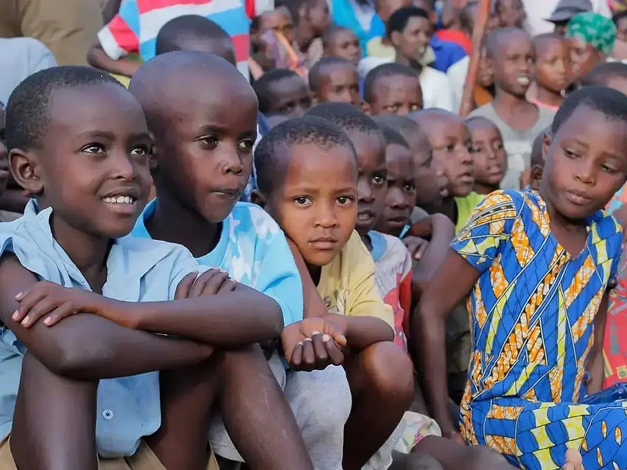 Children sit closely together, watching attentively. Many wear colorful clothing and are surrounded by a large group of other children. The setting appears to be outdoors, with sunlight illuminating their faces.