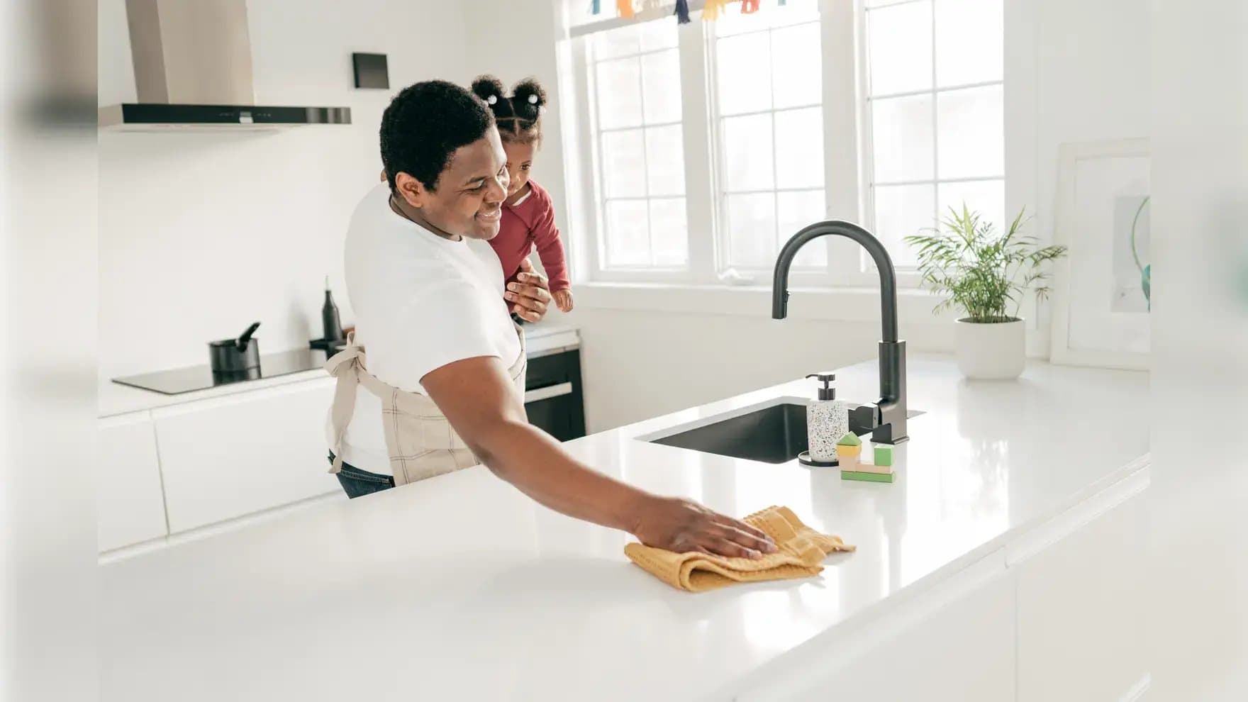 A person cleans a white kitchen counter with a yellow cloth while holding a small child. The bright kitchen has a sink, soap dispenser, potted plant, and large windows.