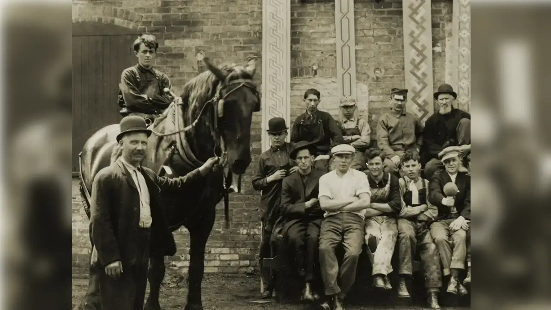 A group of men and a boy sit together, some standing, next to a horse held by a man in a bowler hat. They're in front of a brick building.